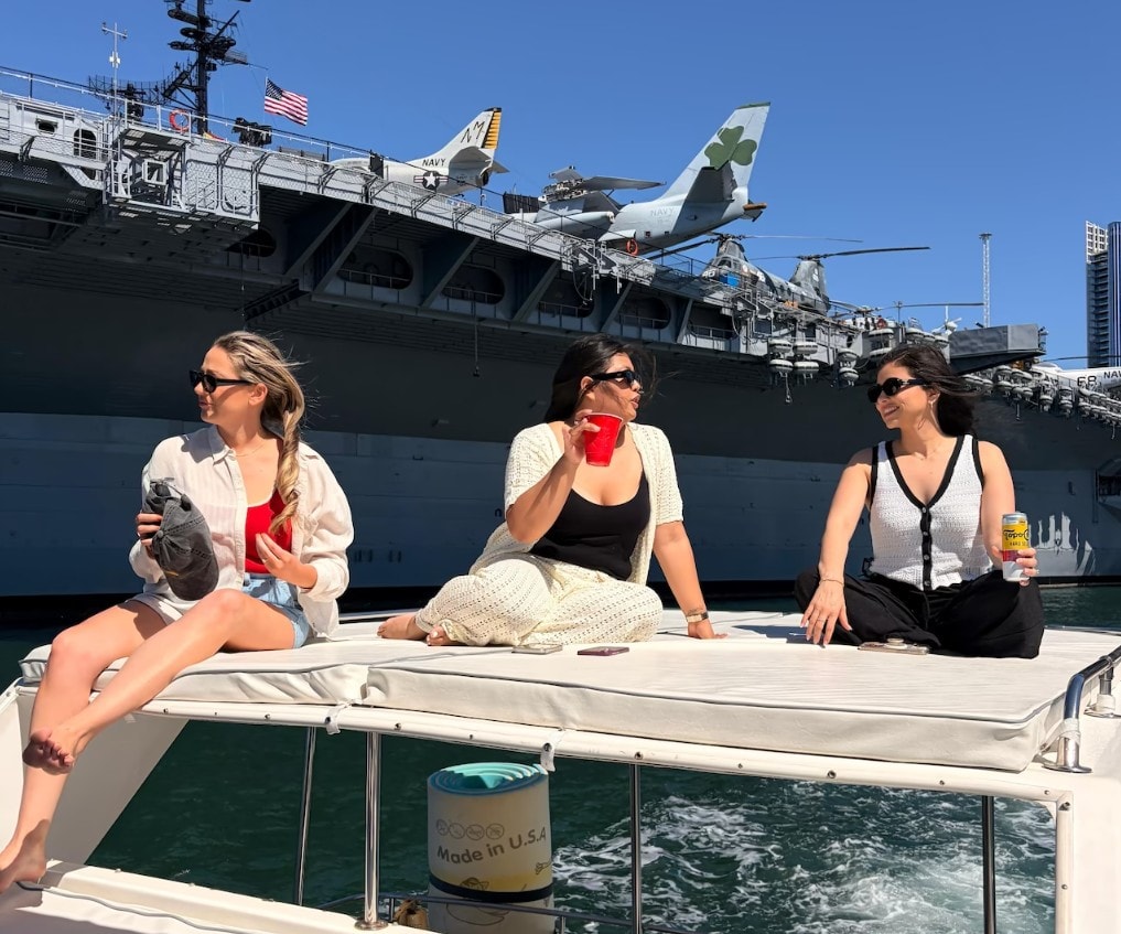Private yacht charter San Diego — three women relaxing on yacht bow with USS Midway aircraft carrier in background