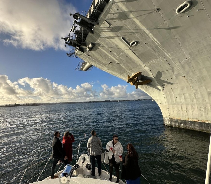 San Diego Bay yacht charter — guests on bow of private party boat directly beneath USS Midway aircraft carrier